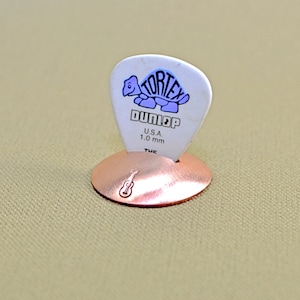 以下が含まれることがあります： ギターピックホルダー、銅色、ギターのデザイン。 白いギターピックには「Tortex DUNLOP U.S.A. 1.0 mm」の文字とカメのグラフィックが描かれています。 ピックの厚さは1.0 mmです。