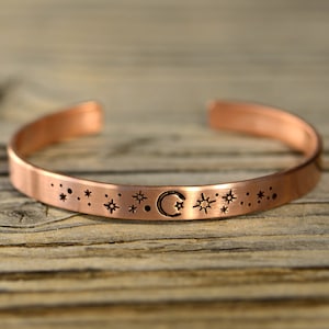 May include: A copper bracelet with a crescent moon and stars design. The bracelet is engraved with a repeating pattern of stars and dots.