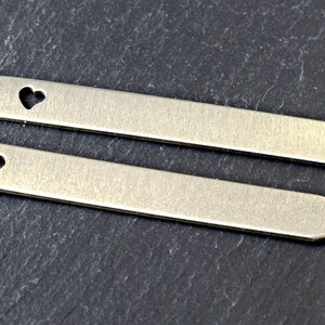 Puede incluir: Dos etiquetas de metal plateado con un corte en forma de coraz&oacute;n. Las etiquetas son rectangulares y tienen un extremo puntiagudo.