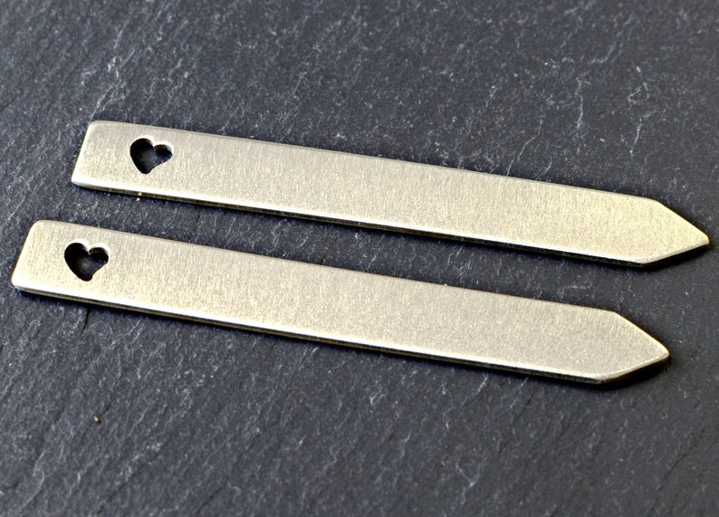 Puede incluir: Dos etiquetas de metal plateado con un corte en forma de coraz&oacute;n. Las etiquetas son rectangulares y tienen un extremo puntiagudo.