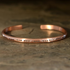 May include: A simple copper bracelet with a hammered finish and a row of small, decorative markings around the band.