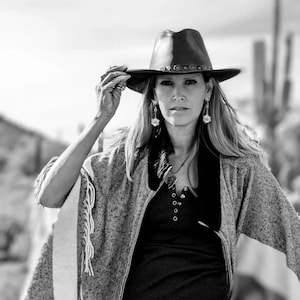 May include: A woman wearing a gray and white fringed poncho, a black shirt, and a brown leather cowboy hat with a decorative band. She is wearing silver earrings and a silver necklace with a heart pendant.