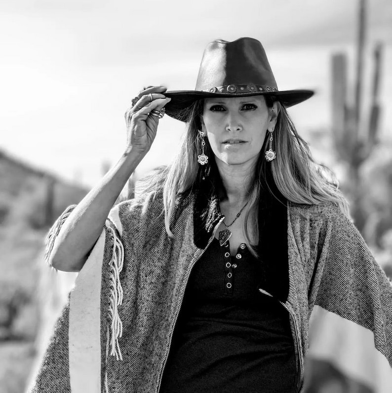 May include: A woman wearing a gray and white fringed poncho, a black shirt, and a brown leather cowboy hat with a decorative band. She is wearing silver earrings and a silver necklace with a heart pendant.