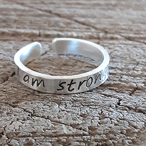Puede incluir: Anillo ajustable de plata con las palabras "Soy fuerte" grabadas en la banda.