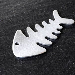 Op de afbeelding: Een zilveren metalen visgraatbedel met een gat om aan een ketting of armband te bevestigen.