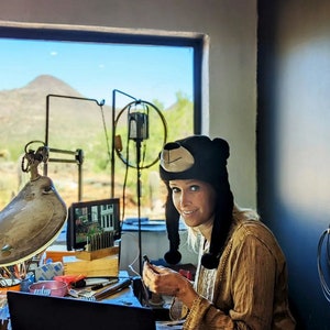 May include: A woman wearing a black bear hat sits at a workbench with a view of a mountain range outside the window. She is working on a jewelry project, using a small tool and a magnifying glass. The workbench is cluttered with tools and supplies.