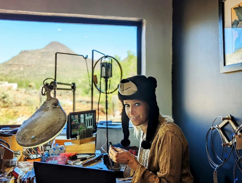 May include: A woman wearing a black bear hat sits at a workbench with a view of a mountain range outside the window. She is working on a jewelry project, using a small tool and a magnifying glass. The workbench is cluttered with tools and supplies.
