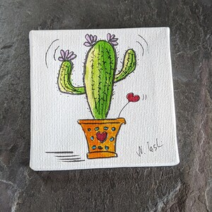 Puede incluir: Un lienzo pintado a mano con un cactus verde con flores rosas en una maceta amarilla con corazones rojos. La firma del artista es visible en la esquina inferior derecha.