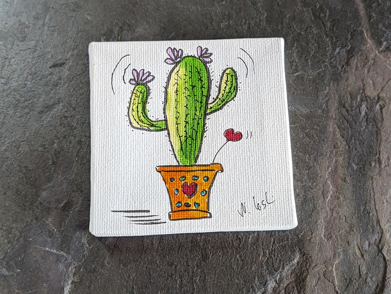 Puede incluir: Un lienzo pintado a mano con un cactus verde con flores rosas en una maceta amarilla con corazones rojos. La firma del artista es visible en la esquina inferior derecha.