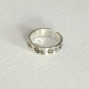 May include: A silver adjustable ring with a black design. The design is a repeating pattern of a stylized letter "G".