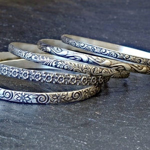 Five silver bangles with intricate floral designs. The bangles are stacked on top of each other, with the most ornate bangle on top.