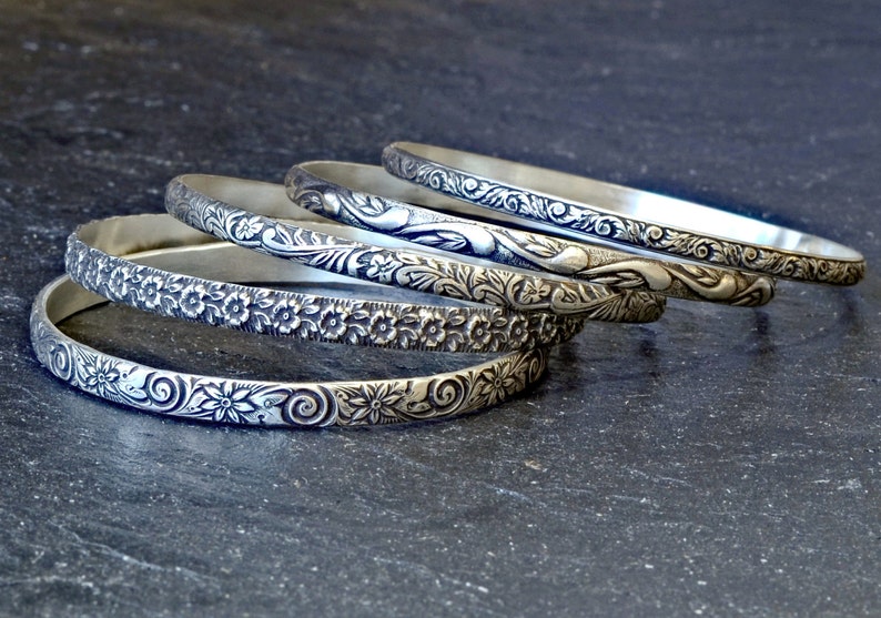 Five silver bangles with intricate floral designs. The bangles are stacked on top of each other, with the most ornate bangle on top.