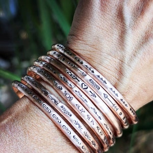 May include: A stack of copper bracelets with various etched designs, including arrows, hearts, and sunbursts.