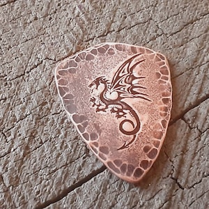 May include: A copper-colored guitar pick with a dragon design. The pick has a textured border and a detailed engraving of a dragon with outstretched wings. The pick is resting on a weathered wooden surface.