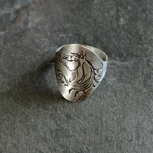 May include: A silver ring with a detailed design of a mythical creature, possibly a griffin, etched into the surface.