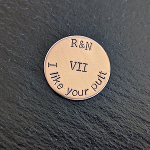 May include: A round, copper-colored golf ball marker with the text "R&N VII I like your putt" engraved on it.