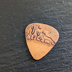 May include: A copper guitar pick with a design of a coyote howling at the moon in a desert landscape with cacti.