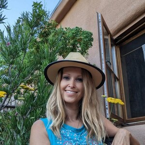 May include: A woman with long blonde hair is wearing a blue and red patterned tank top and a wide-brimmed straw hat with a black band. She is smiling and looking at the camera.