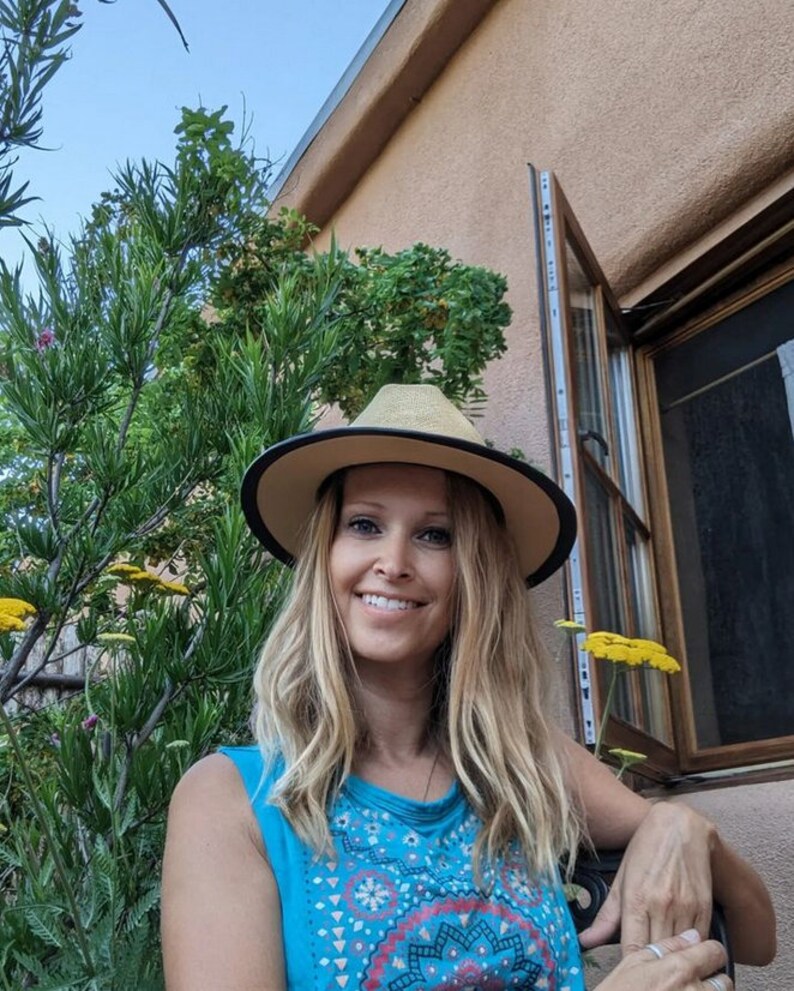 May include: A woman with long blonde hair is wearing a blue and red patterned tank top and a wide-brimmed straw hat with a black band. She is smiling and looking at the camera.