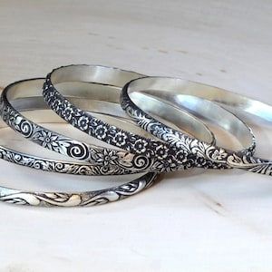 May include: Four silver bangles with intricate floral designs. The bangles are stacked on top of each other, with the most ornate design on the bottom.