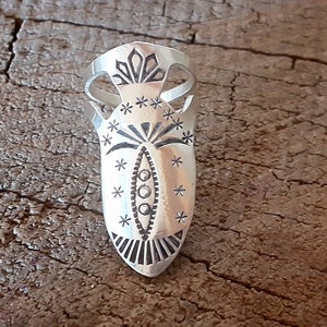 May include: A silver-colored ring with an elongated, shield-like design. The ring features intricate etched patterns, including star-like shapes and a central oval with three dots. The ring's design is reminiscent of Native American jewelry.