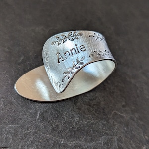 May include: A silver-colored ring with the name "Annie" engraved on it. The ring has a leaf design around the edges and is set on a silver oval base. The ring is made of metal and has a brushed finish.