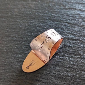 Puede incluir: Una púa de guitarra de cobre con el texto "March 17" y una pequeña ilustración de guitarra. La púa tiene un diseño curvado y está hecha de un material metálico brillante. La púa está diseñada para ser usada en un dedo.