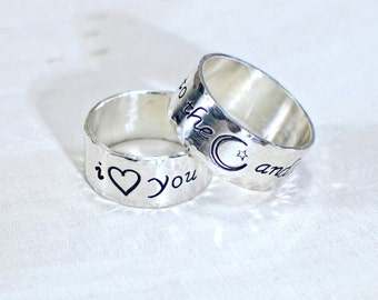 May include: Two silver rings with hammered textures. One ring has the words "i heart you" engraved, and the other has "to the moon and" with a moon and star symbol. The rings are likely for couples.
