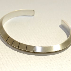 May include: A silver bangle bracelet with a series of rectangular cutouts along the bottom edge.