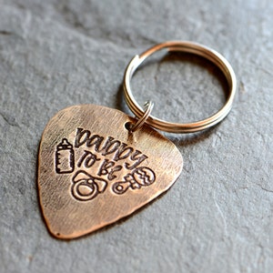 May include: A bronze-colored guitar pick keychain with the text "Happy to be Dad" engraved on it. The keychain also features a small engraved image of a baby bottle, a rattle, and a pacifier.