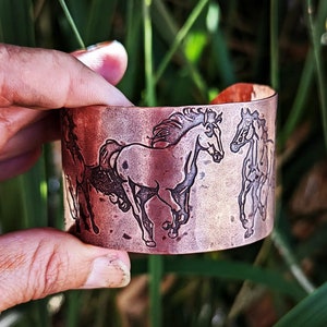 May include: Copper cuff bracelet featuring etched running horses. The wide, hammered-finish bracelet has a rustic, vintage look. The horses are depicted in a detailed, stylized manner, adding a touch of Western charm. The bracelet is approximately 2 inches wide.