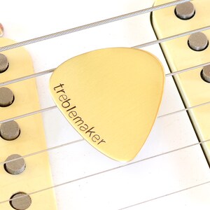 May include: Close-up of a guitar with strings and a cream-colored pickguard. A gold-colored guitar pick with the word "treblemaker" is positioned between the strings. The pickguard has three visible pickups.