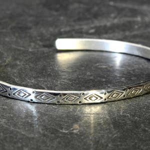 May include: A silver bangle bracelet with a geometric pattern. The bracelet is made of a thin band of metal and has a simple, elegant design.