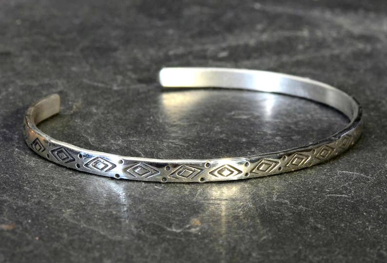 May include: A silver bangle bracelet with a geometric pattern. The bracelet is made of a thin band of metal and has a simple, elegant design.