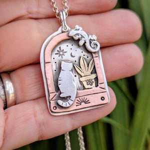 Può includere: Collana con pendente in argento raffigurante un gatto, una pianta e una lucertola all'interno di una cornice a finestra. Il pendente ha dettagli in rame e oro, con uno sfondo di cielo stellato. La catena della collana è d'argento.