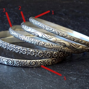 May include: Five silver bangles with different floral designs. The bangles are stacked on top of each other.