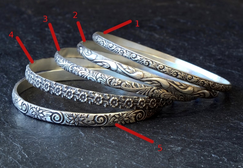 May include: Five silver bangles with different floral designs. The bangles are stacked on top of each other.