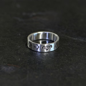 May include: A silver band ring with a design of four small circles and a bird in the center.
