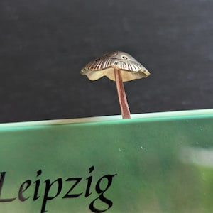 May include: A small, detailed mushroom sculpture with a textured cap and a brown stem. The mushroom is made of a metallic material and is set against a dark background. The word "Leipzig" is partially visible in the foreground.