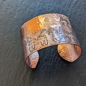May include: A wide copper cuff bracelet with a hammered texture and a running horse design. The bracelet has a rustic, handcrafted appearance and is open at the bottom. The cuff is approximately 2 inches wide.