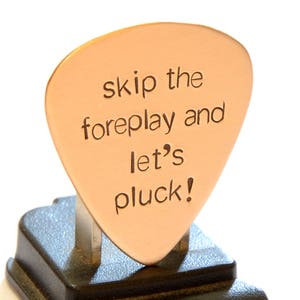 May include: A copper guitar pick with the text "skip the foreplay and let's pluck!" engraved on it. The pick is displayed on a black stand.