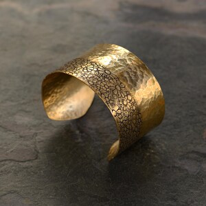 May include: A wide, hammered brass cuff bracelet. The bracelet has a floral pattern around the top edge. The cuff is approximately 2 inches wide.
