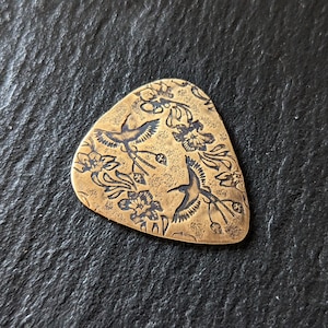 Pode incluir: Um palheta de guitarra dourada com um design floral e pássaros em voo. A palheta é feita de metal e tem uma superfície texturizada.