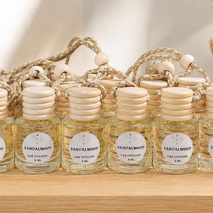 Peut inclure: Une collection de petits flacons diffuseurs de voiture en verre transparent avec des bouchons à vis en bois clair et des cordons de suspension. Chaque flacon contient un liquide jaune pâle et porte une étiquette ronde blanche avec le texte SANDALWOOD CAR DIFFUSER 8 ML.