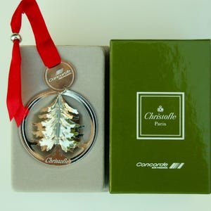 Könnte beinhalten: Silberner Weihnachtsbaumschmuck mit rotem Band und silberner Kugel. Der Schmuck befindet sich in einem silbernen Kreis und neben einer grünen Schachtel mit den Aufschriften "Christofle Paris" und "Concorde Air France".
