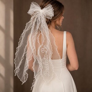 Puede incluir: Un velo de tul blanco con un lazo grande en la parte superior y bordes con volantes. El velo está bordado con el texto "Bride to Be" en letras cursivas blancas. La tela transparente cae en cascada creando un efecto de capas.