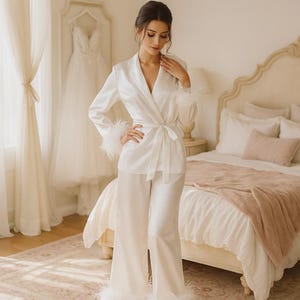 May include: A white satin pajama set with feather trim. The set includes a wrap-style jacket with a tie waist and wide-leg trousers. A wedding dress hangs in the background. The bedroom has a bed with white linens and a pink throw.