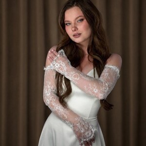Puede incluir: Un vestido de novia blanco con delicadas mangas de encaje. Las mangas largas y ajustadas presentan intrincados dise&ntilde;os florales. El vestido tiene un dise&ntilde;o simple y elegante. El fondo es marr&oacute;n neutro.