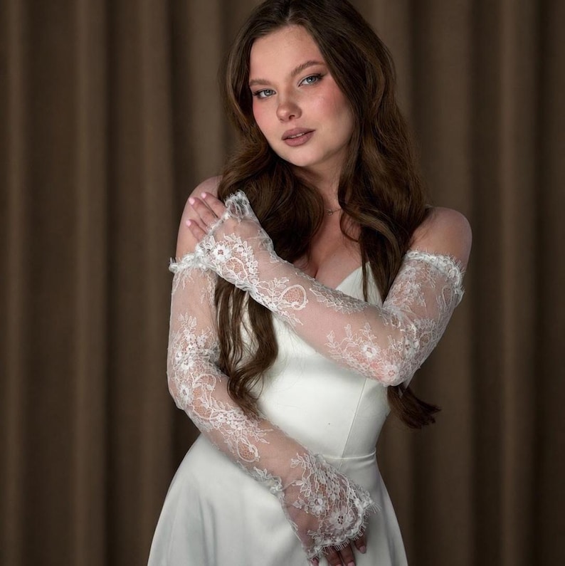 Puede incluir: Un vestido de novia blanco con delicadas mangas de encaje. Las mangas largas y ajustadas presentan intrincados dise&ntilde;os florales. El vestido tiene un dise&ntilde;o simple y elegante. El fondo es marr&oacute;n neutro.