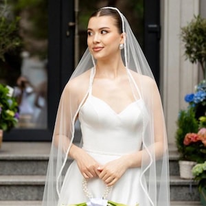 Puede incluir: Un velo de novia blanco con un delicado borde de cuentas a lo largo de los contornos, usado sobre un vestido blanco sin mangas con escote corazón. La tela de tul transparente cae sobre los hombros, complementada con una diadema de cuentas.
